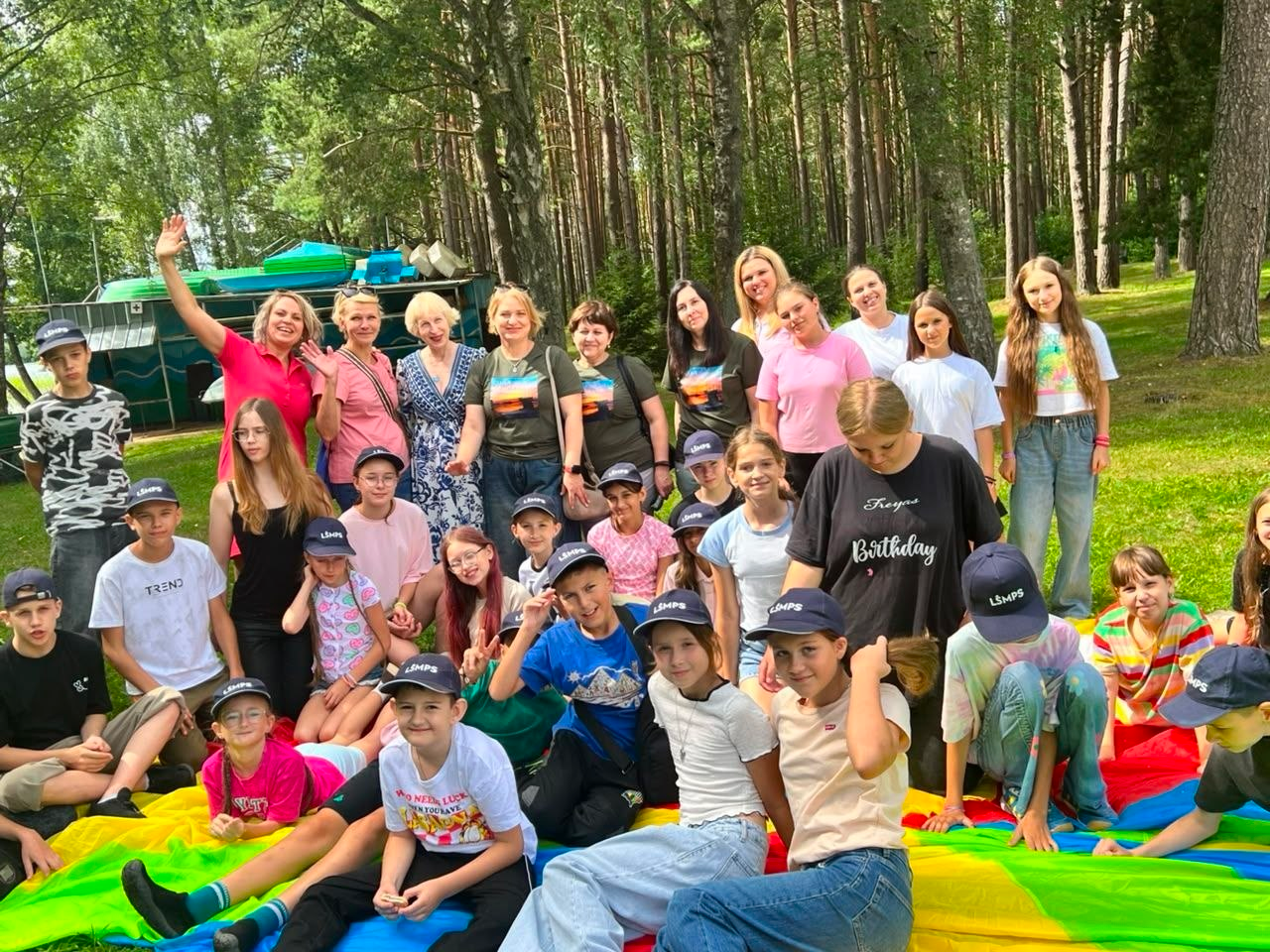 Делегація херсонських освітян та діти з України під час візиту до литовського міста-побратима Висагінаса. Велика група українських дітей у фірмових синіх кепках литовської профспілки освітян «LŠMPS» відпочиває в літньому таборі на природі. Позаду них стоять дорослі — представники Голопристанської громади та херсонські педагоги, які відвідали Литву в межах обміну досвідом та зміцнення міжнародного партнерства.