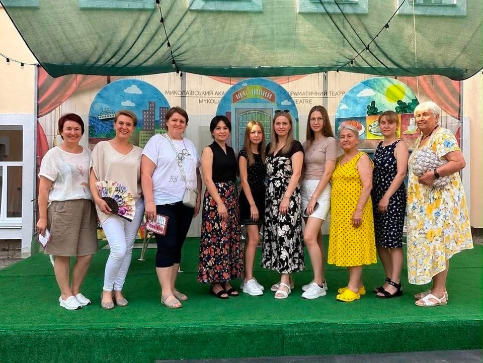 ВПО з Херсонщини відвідали арттерапевтичну виставу в Миколаївському академічному художньому драматичному театрі. Група херсонок, які тимчасово мешкають у Миколаєві, позує для спільного фото на літній сцені театру після перегляду вистави «Slow twisting на чотири голоси». Десять учасниць стоять на тлі театральних декорацій із написом «Щасливий».