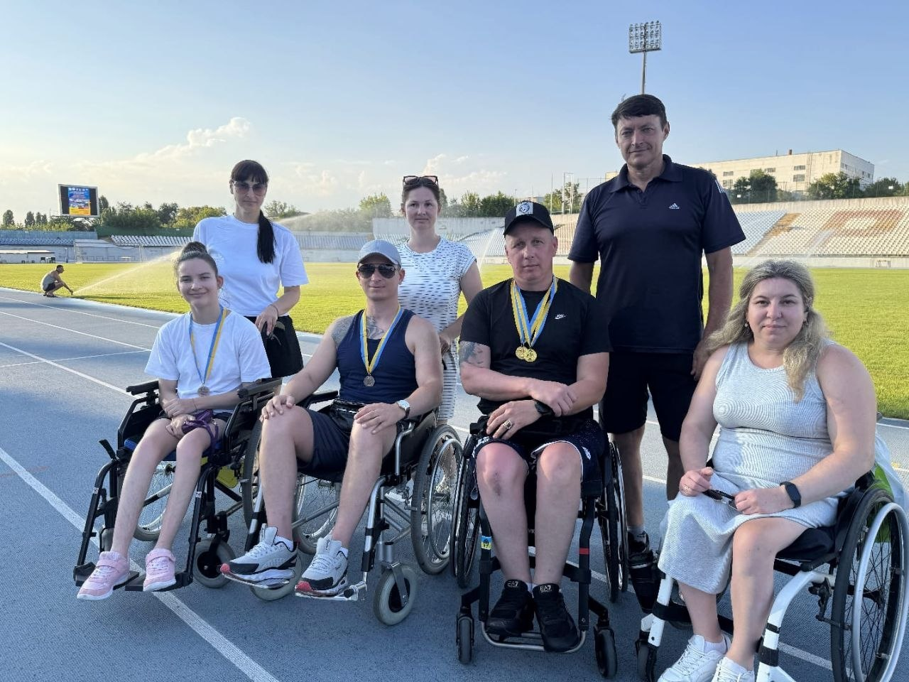 Параатлети з Херсонщини зі своїми тренерами на чемпіонаті України з паралегкої атлетики у Кропивницькому. На синій біговій доріжці стадіону в кріслах колісних сидять призери змагань з медалями: Анастасія Клименко, Антон Саітов, Денис Горбенко та Наталія Сівак. За ними стоять тренери та супроводжуючі, серед яких Олексій Тімолаєв, святкуючи завоювання 18 нагород для області.