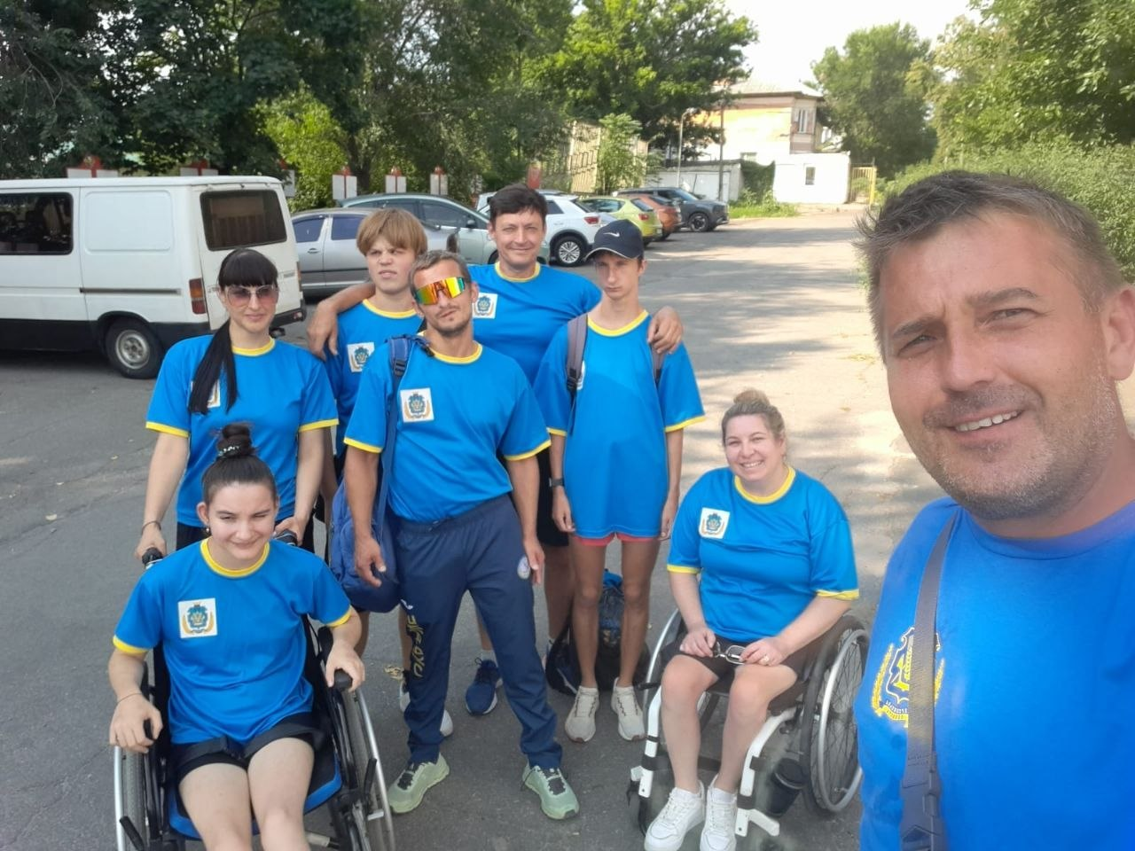 Спільне селфі збірної команди параатлетів Херсонщини та їхніх супроводжуючих на чемпіонаті України у Кропивницькому. Чоловік на передньому плані робить групове фото, на якому дев'ять учасників у фірмових синьо-жовтих спортивних футболках із гербом області посміхаються на тлі відкритого майданчика та припаркованих автомобілів. Серед спортсменів — дві дівчини у кріслах колісних.