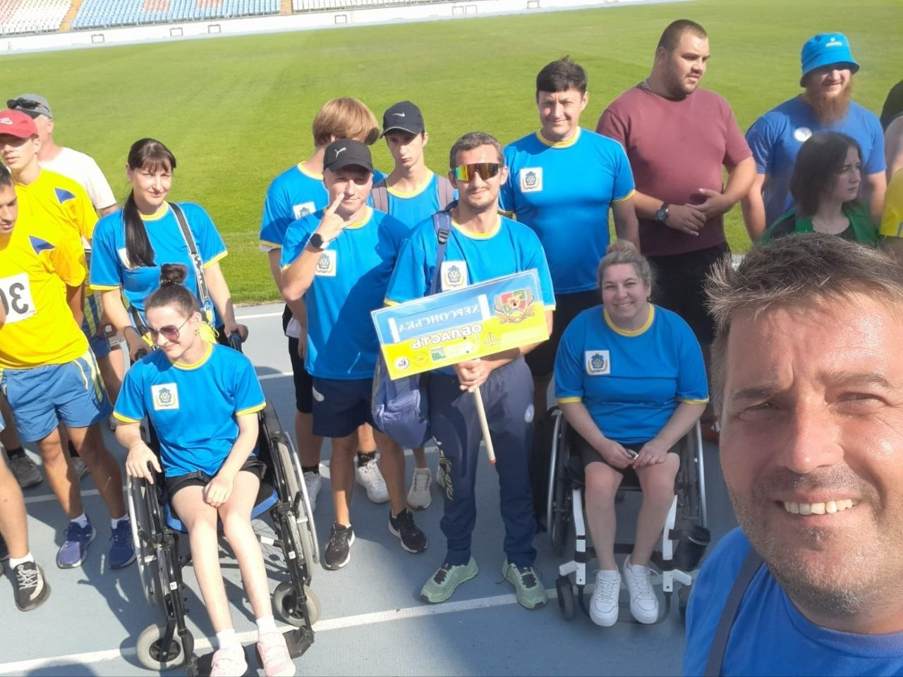 Збірна команда параатлетів Херсонщини з табличкою області на чемпіонаті України у Кропивницькому. Чоловік на передньому плані робить велике групове селфі спортсменів та тренерів у синьо-жовтих фірмових футболках на синій біговій доріжці стадіону. У центрі кадру один з атлетів тримає офіційну жовто-блакитну табличку з гербом та написом «Херсонська область».
