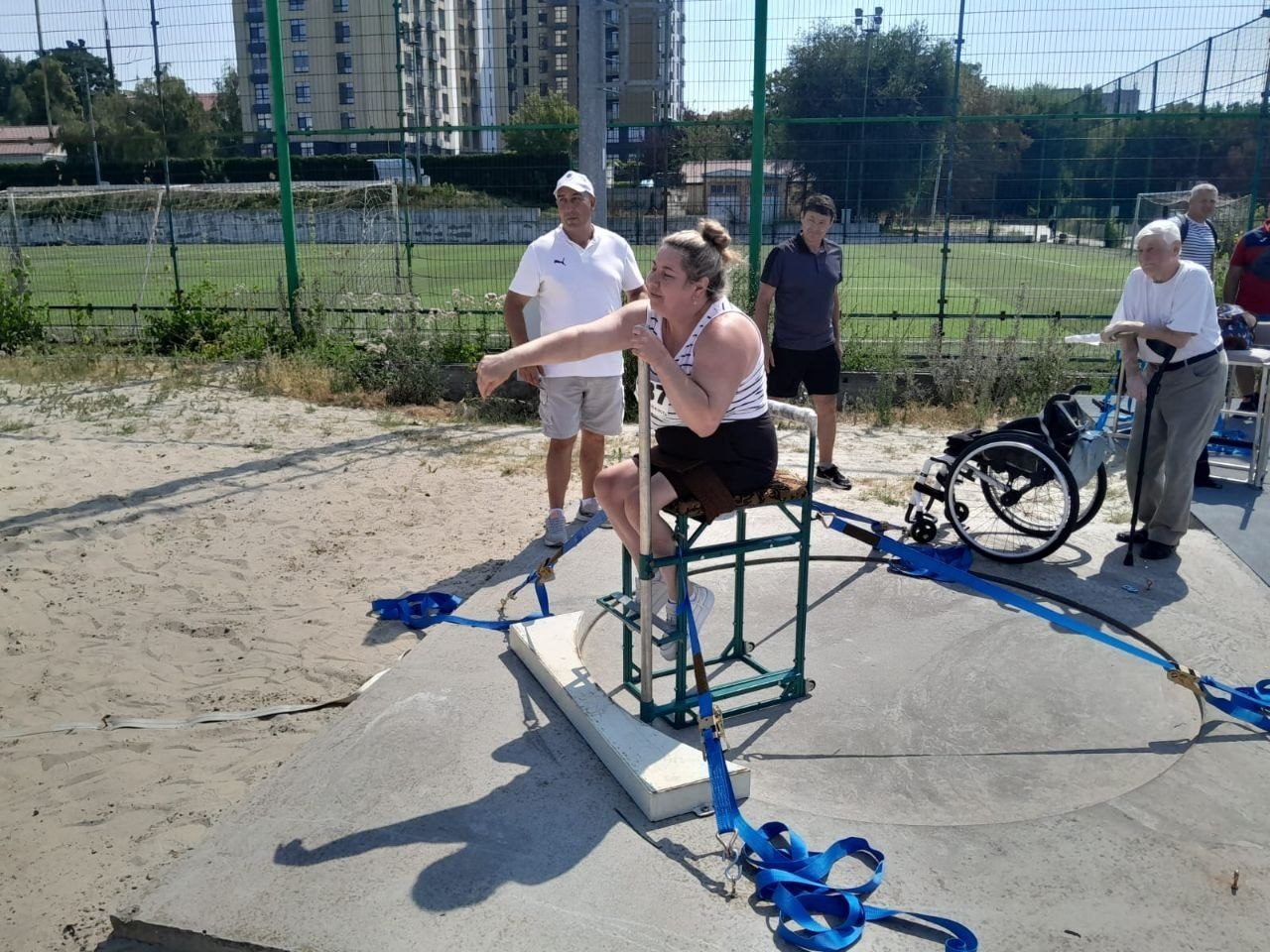 Параатлетка Наталія Сівак виконує поштовх ядра на чемпіонаті України з паралегкої атлетики. Спортсменка зафіксована у момент кидка, сидячи на спеціальному металевому станку для метання, який закріплений ременями на бетонному секторі стадіону. За її виступом уважно спостерігають судді, тренери та інші учасники змагань.