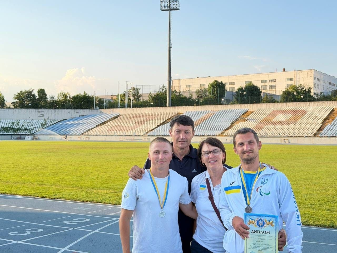 Українські параатлети Артем Мудрак та Іван Тетюхін із тренерами на стадіоні у Кропивницькому. Спортсмени у фірмовій спортивній формі з медалями на синьо-жовтих стрічках позують на біговій доріжці, тримаючи офіційний диплом змагань. Поруч із ними стоять їхні тренери Наталя Туришева та Олексій Тімолаєв на тлі трибун та зеленого поля.