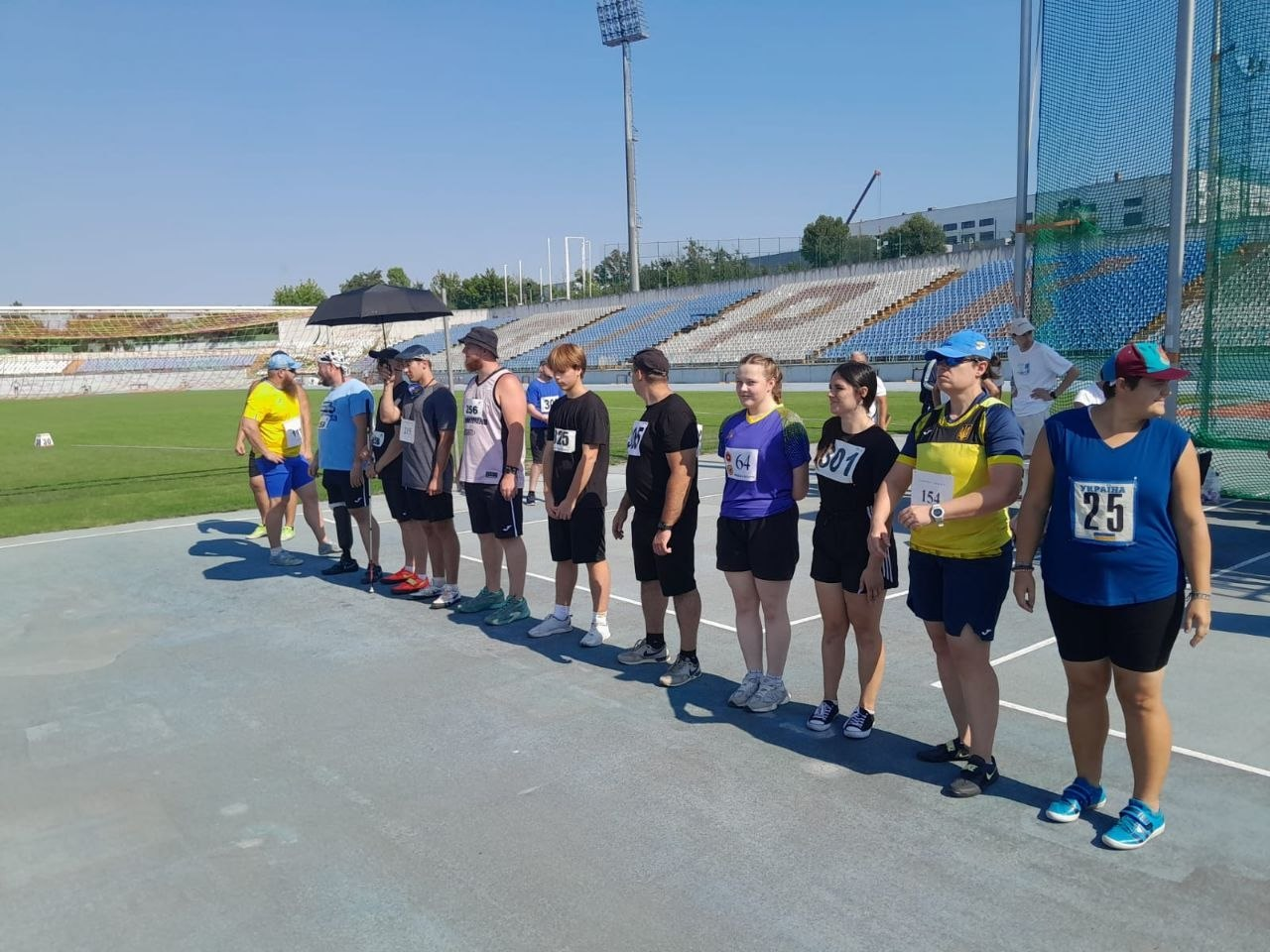 Параатлети вишикувалися на стадіоні перед початком змагань або церемонією нагородження на чемпіонаті України. Група спортсменів із номерами на спортивній формі стоїть в один ряд на синій біговій доріжці. Позаду них видно трибуни стадіону, зелене футбольне поле та сітку сектору для метань під ясним небом у Кропивницькому.