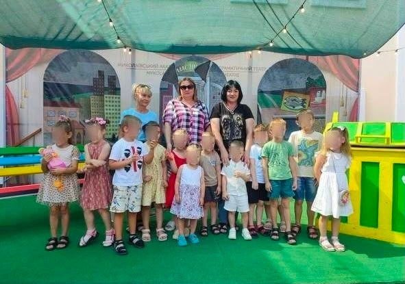 Група юних переселенців із Херсонщини разом із трьома дорослими жінками позує для фото на зеленій сцені у літньому дворику Миколаївського академічного художнього драматичного театру. Цей візит організував Координаційний центр «Вільні разом». Діти різного віку одягнені в літній одяг, дехто тримає іграшки. Позаду них встановлено театральні декорації із зображенням міських будинків, а зверху натягнуто зелений захисний навіс із гірляндами. Світлина зроблена після вистави «Пес Тимко рятує друзів».