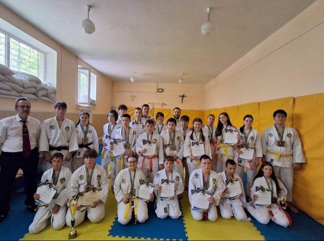 Група юних спортсменів різного віку у кімоно позує з медалями, грамотами та кубком на татамі після завершення відкритого Кубка Херсонської області з рукопашного бою пам’яті загиблого військовослужбовця Дмитра Чупикова. Позаду та збоку від дітей стоять троє дорослих тренерів та суддів. Учасники змагань тримають у руках завойовані нагороди, демонструючи результат своїх виступів у розділах легкого, повного контакту, самозахисту та ката.