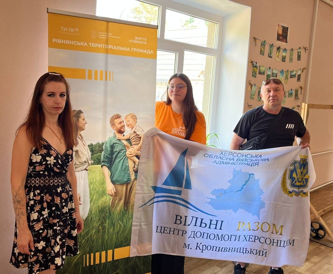 Дві жінки та чоловік — фахівці координаційного центру «Вільні разом» — позують у приміщенні біля банера Рівнянської територіальної громади. Вони тримають у руках прапор Центру допомоги херсонцям у місті Кропивницький із написом «Херсонська обласна військова адміністрація». Світлина зроблена під час виїзної зустрічі у Центрі життєстійкості з родинами новоприбулих переселенців із Херсонщини.