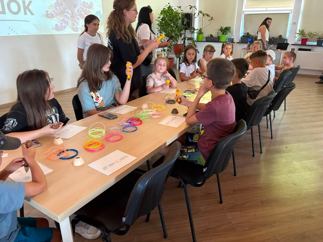 Юні переселенці з Херсонщини беруть участь у тематичних іграх та творчому майстер-класі за довгим столом у Кропивницькому. Фахівчиня «Майстерні 3D-іграшок» показує дітям елементи для моделювання, а на столі перед ними розкладено мотки різнокольорового пластику та заготовки для створення фігурок за допомогою 3D-ручок. Захід присвячено Дню молоді та організовано спільно з Верхньорогачицькою СВА.
