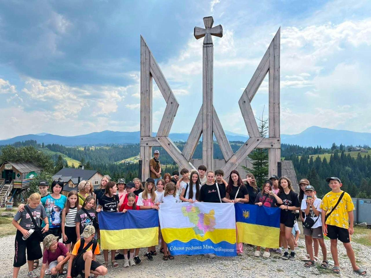 Група дітей загиблих і зниклих безвісти Захисників та Захисниць України з Херсонщини на відпочинку в Яремче в межах проєкту «Живі Серцем». Діти стоять із прапорами біля великого дерев’яного Тризуба на тлі Карпатських гір перед початком тижневої екскурсійної та розважальної програми.