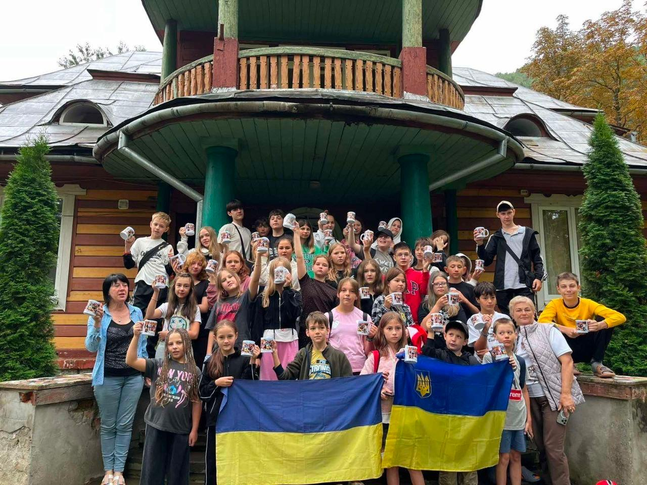 Велика група дітей та підлітків разом із дорослими супроводжуючими позирує для спільного фото на сходах колоритного дерев'яного будинку в Яремче. Це діти загиблих (померлих) Захисників і Захисниць України, а також діти з родин зниклих безвісти за особливих обставин із Херсонщини, які приїхали на відпочинок у межах соціально-освітнього проєкту «Живі Серцем».
