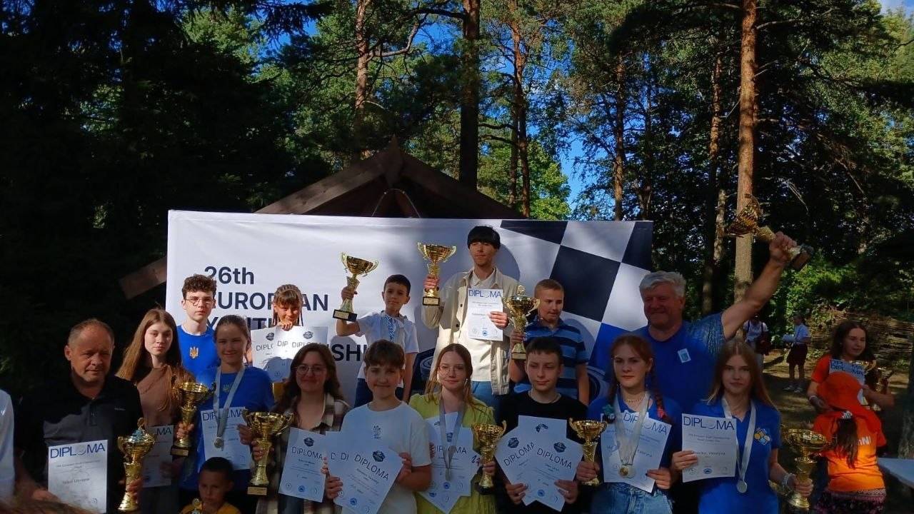 Збірна України з шашок на церемонії нагородження Молодіжного Чемпіонату Європи в Таллінні. Велика група молодих спортсменів, серед яких «срібний» призер Артем Філенко, разом із тренерами позирують на тлі офіційного банера змагань під відкритим небом, тримаючи в руках завойовані кубки, дипломи та медалі.