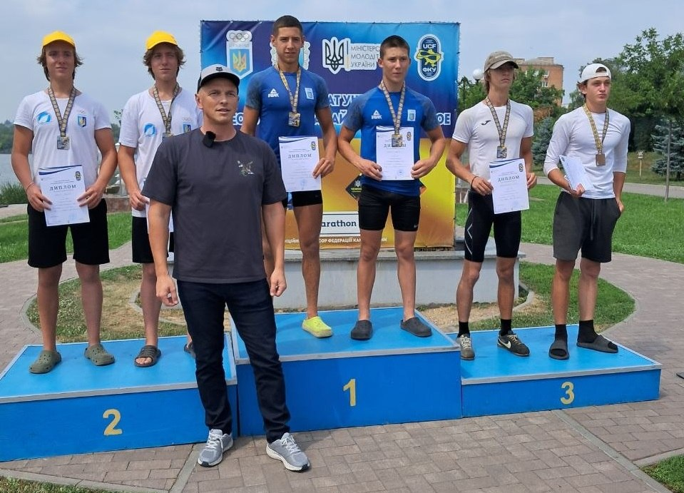 Церемонія нагородження призерів Чемпіонату України з веслування на байдарках і каное. Три пари юних спортсменів з медалями та дипломами в руках стоять на трирівневому синьому п'єдесталі пошани на тлі офіційного банера Міністерства молоді та спорту України та Федерації каное України. Попереду, ліворуч від першого місця, стоїть чоловік у темному одязі та кепці (тренер або організатор).