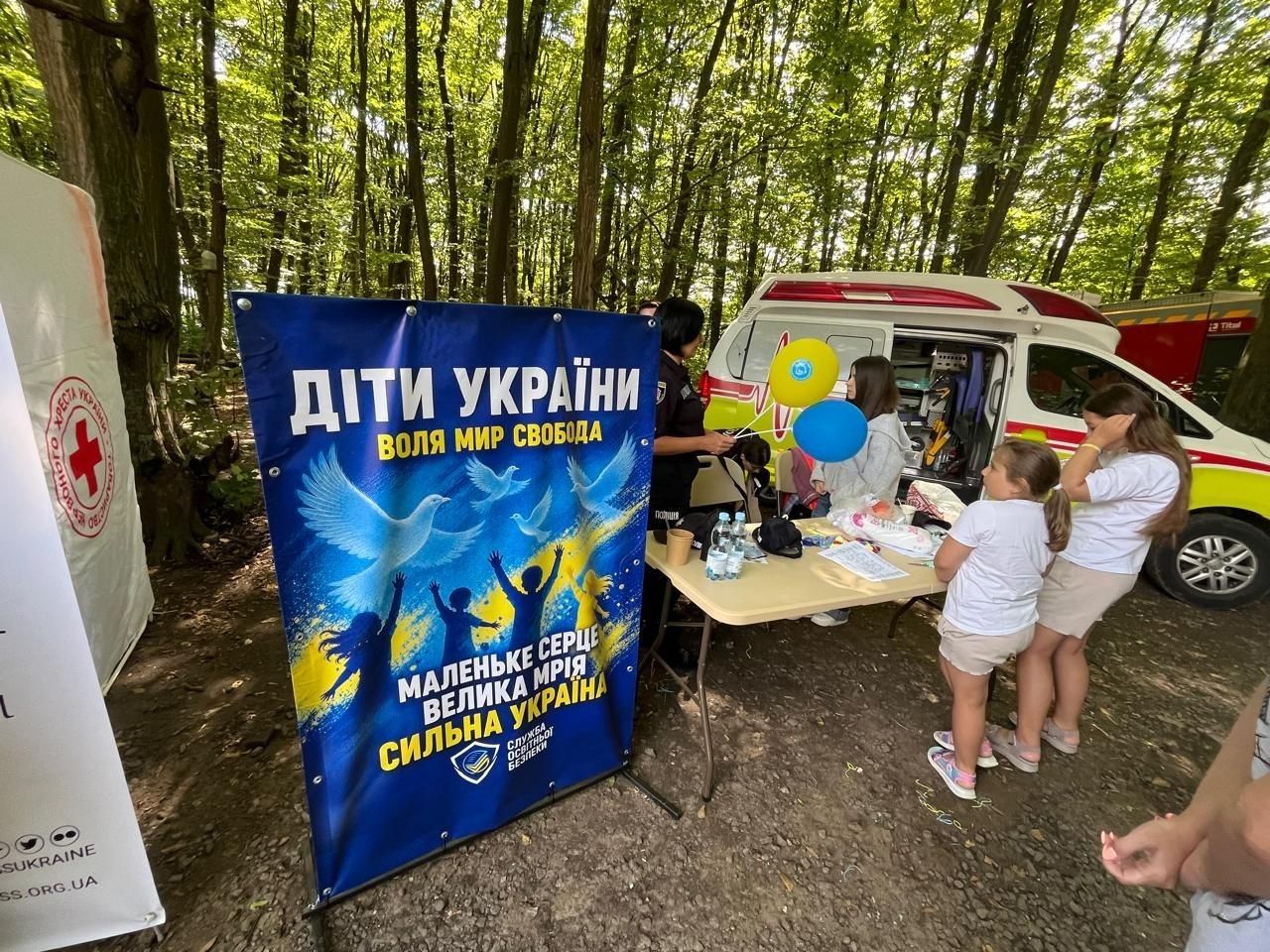 Переселенці з Херсонщини та представники координаційного центру «Вільні разом» на спортивному фестивалі «Воля Fest» у лісовій зоні Хмельницького. На передньому плані — великий синій банер заходу з написами «ДІТИ УКРАЇНИ» та «МАЛЕНЬКЕ СЕРЦЕ ВЕЛИКА МРІЯ СИЛЬНА УКРАЇНА». Поруч розташована локація Товариства Червоного Хреста України та автомобіль екстреної допомоги для супроводу змагань. Біля столу з водою та матеріалами для творчого майстер-класу стоять діти з патріотичними жовто-синіми кульками та організатори події.