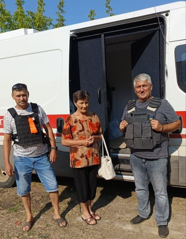 Волонтери Юрій Раков та Анатолій Лелюх у бронежилетах стоять біля евакуаційного мікроавтобуса разом із літньою жінкою, яку вони евакуювали з села Раківка Бериславської громади.