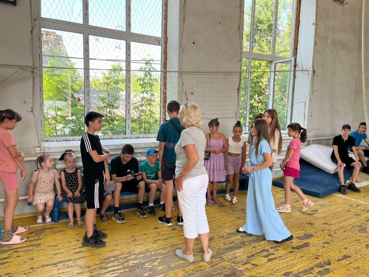 Діти різного віку та викладачі збираються у великому спортивному залі Калинівської громади на відкриття творчого простору від Херсонського обласного палацу культури. Частина дітей сидить на лавах біля великих вікон, інші спілкуються у центрі зали, готуючись до занять із хореографії та акторської майстерності.