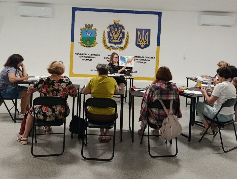 Ведуча проводить заняття з англійської мови для переселенок із Херсонщини в координаційному центрі «Вільні разом» у Черкасах. Жінки сидять півколом за столами й обговорюють навчальні матеріали з історії США та феміністичного руху, а на стіні за ведучою розміщено банер із символікою Чаплинської та Асканіатівської громад.