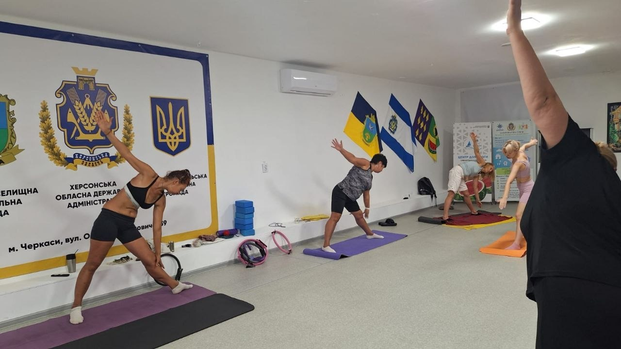 Переселенки з Херсонщини виконують вправи на килимках під час заняття з йоги та стретчингу в координаційному центрі «Вільні разом» у Черкасах. На стінах зали розміщено великий банер Херсонської ОВА та прапори громад.