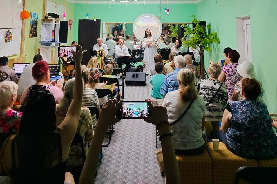 Глядачі різного віку у просторі «Дитинство» ГО «Сильні бо вільні» захоплено дивляться концерт артистів Херсонського театру імені Миколи Куліша та знімають виступ на смартфони. Подія відбувається у межах проєкту БФ «Крила Фундейшн» за підтримки Посольства Естонії в Україні, а на задньому плані над сценою світиться кругла проєкція-банер програми «Коло життя. Літо».
