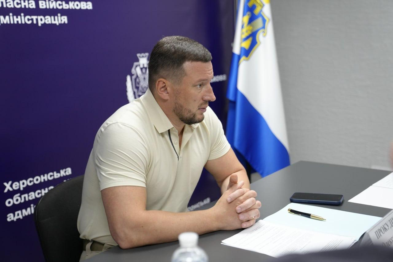 Начальник Херсонської ОВА Олександр Прокудін проводить засідання штабу з оперативного реагування на наслідки російських ударів по енергетиці міста. Він сидить за робочим столом із документами на тлі офіційного банера адміністрації та прапора України.