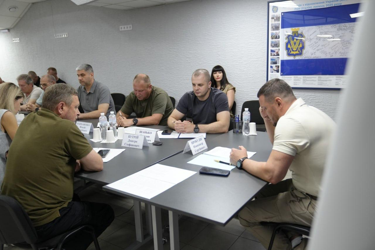 Начальник Херсонської ОВА Олександр Прокудін у світлій футболці-поло сидить праворуч за робочим столом із документами та проводить засідання оперативного штабу. Навпроти нього ліворуч сидить перший заступник начальника ОВА Дмитро Бутрій у зеленій футболці-поло, за яким далі сидить заступниця начальника ОВА Ольга Малярчук у світлій блузці, на протилежному боці столу присутній начальник Херсонської МВА Ярослав Шанько у темно-синій футболці, а також інші члени засідання, які сидять далі вздовж столу.
