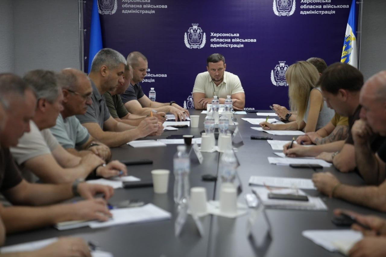 Начальник Херсонської ОВА Олександр Прокудін у світлій футболці-поло сидить у центрі на чолі столу та проводить засідання штабу з оперативного реагування на наслідки російських ударів по енергетиці на тлі фіолетового офіційного банера. Обабіч нього розташовані інші члени засідання, де праворуч від нього сидять перший заступник начальника ОВА Дмитро Бутрій у зеленій футболці-поло, заступниця начальника ОВА Ольга Малярчук у світлій блузці та заступник начальника ОВА Євгеній Ігнатенко в хакі-футболці, яких частково видно у кадрі, а також директор Департаменту цивільного захисту Євген Пісоцький у темній футболці; ліворуч за столом сидить начальник Херсонської МВА Ярослав Шанько у темно-синій футболці.
