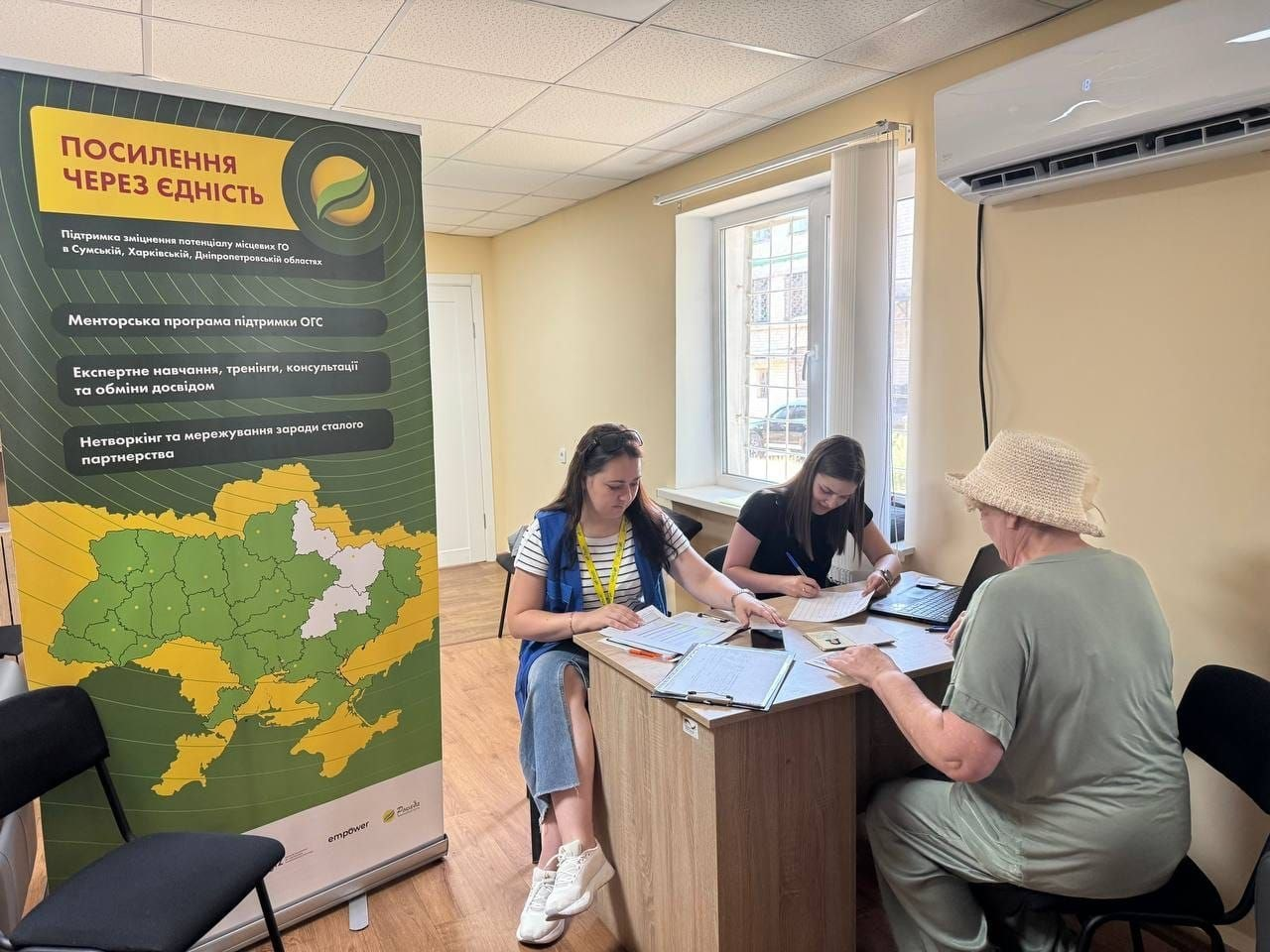 Представниці БФ «Рокада» та координаційного центру «Вільні разом» у Дніпрі приймають заяви й консультують переселенку з Херсонщини у Жовтих Водах. Поруч на передньому плані встановлено великий інформаційний банер проєкту «Посилення через єдність» із картою Сумської, Харківської та Дніпропетровської областей.