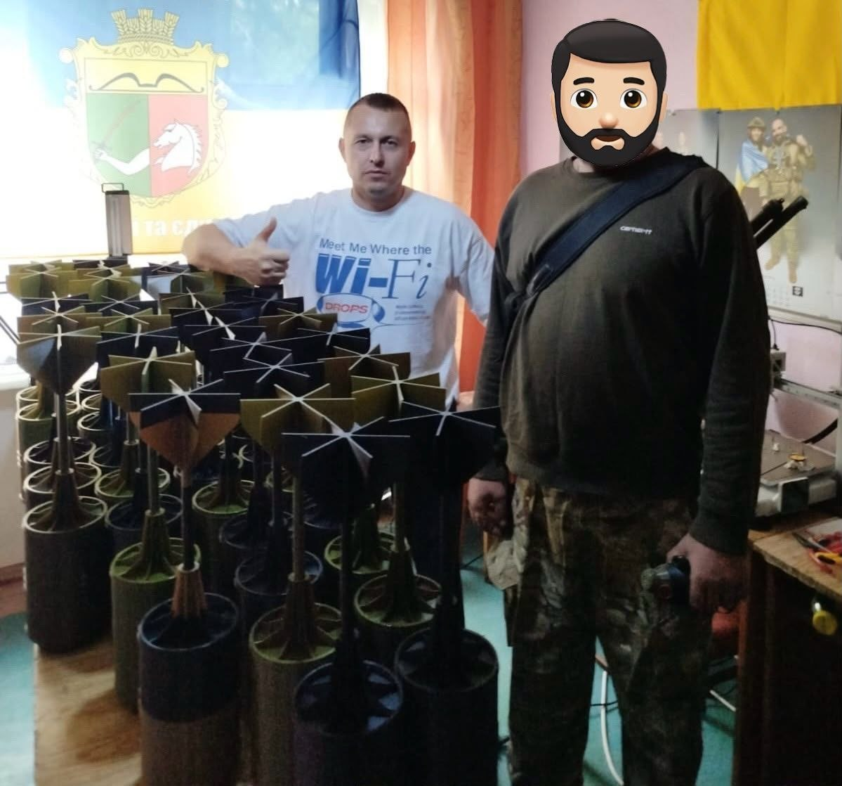 Представник Чулаківської СВА у білій футболці показує великий палець угору, позуючи разом із військовослужбовцем із закритим обличчям на тлі прапора України та прапора Скадовського району з гербом, поруч із великою кількістю виготовлених та переданих на фронт хвостовиків-стабілізаторів для БПЛА.