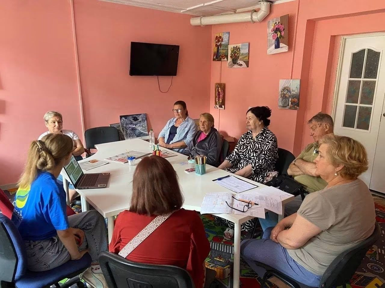 Фахівці БФ «Рокада» проводять інформаційну зустріч із медіаграмотності для переселенців із Херсонщини в координаційному центрі «Вільні разом» у Львові. Учасники заходу сидять за великим білим столом у рожевій кімнаті перед ноутбуком та документами, слухаючи ведучу в синій брендованій футболці.