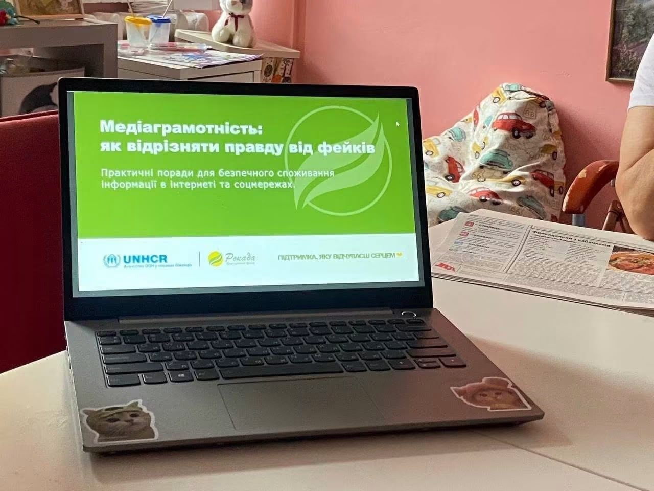 Відкритий ноутбук стоїть на білому столі під час заходу в координаційному центрі «Вільні разом» у Львові. На його зеленому екрані відображається презентація «Медіаграмотність: як відрізняти правду від фейків» із логотипами UNHCR та БФ «Рокада», а поруч лежать друковані інформаційні матеріали.