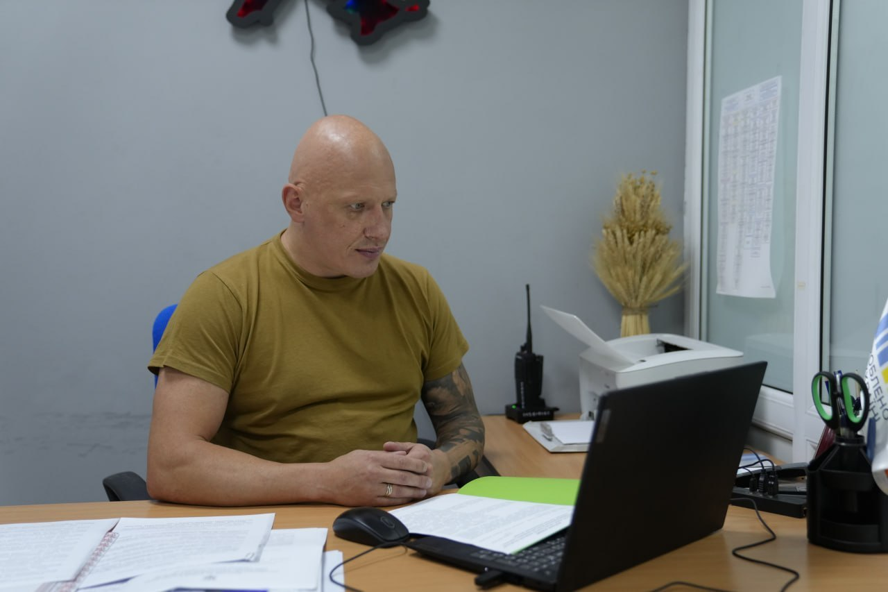 Заступник начальника Херсонської ОВА Євгеній Ігнатенко в оливковій футболці сидить за робочим столом перед ноутбуком із документами та проводить засідання обласної комісії з питань надання одноразової матеріальної допомоги жителям Херсонської області, на задньому плані в кабінеті стоять рація та декоративний дідух із колосків.