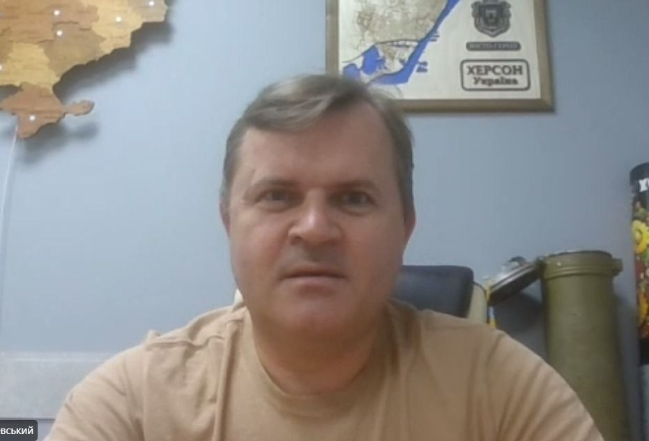 Заступник начальника Херсонської ОВА Володимир Клюцевський у світло-коричневій футболці проводить засідання Гуманітарного штабу в режимі відеоконференції. На стіні за ним розміщено дерев'яну карту України, плакат із написом «Херсон. Україна. Місто-герой» та відстріляну гільзу артилерійського снаряда.