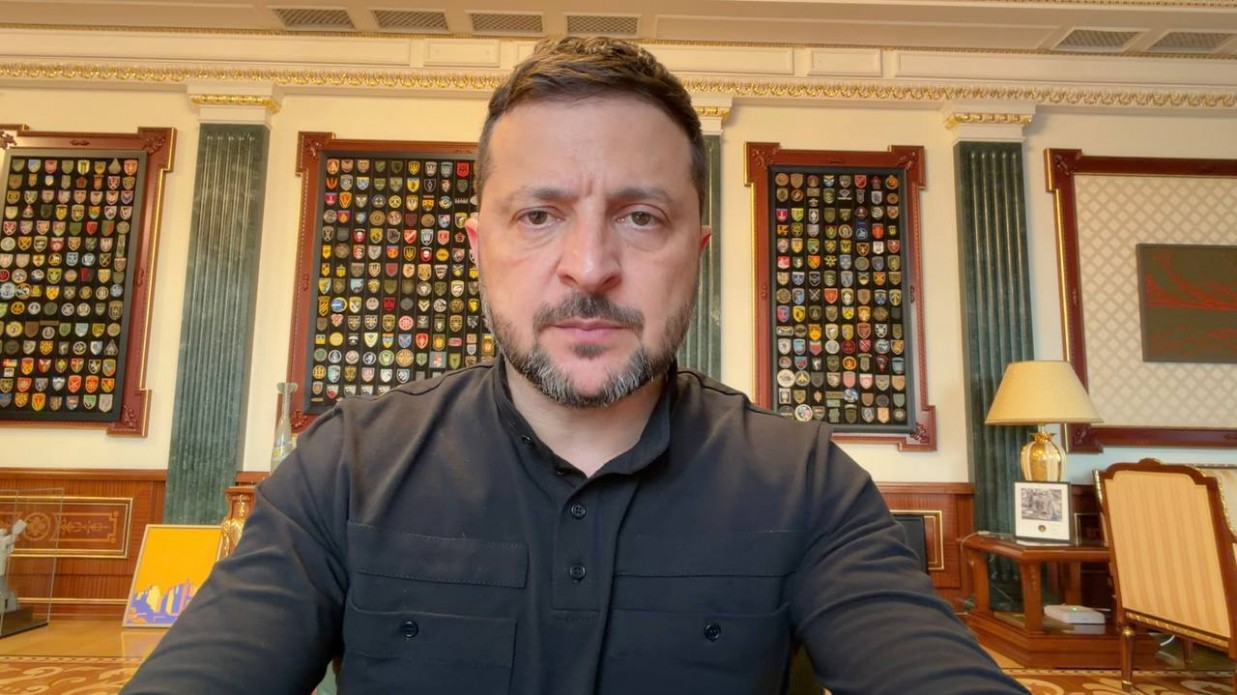 Президент України Володимир Зеленський у чорній сорочці-поло виступає з офіційним вечірнім відеозверненням на тлі стін робочого кабінету, прикрашених великими колекціями військових шевронів у дерев'яних рамках.