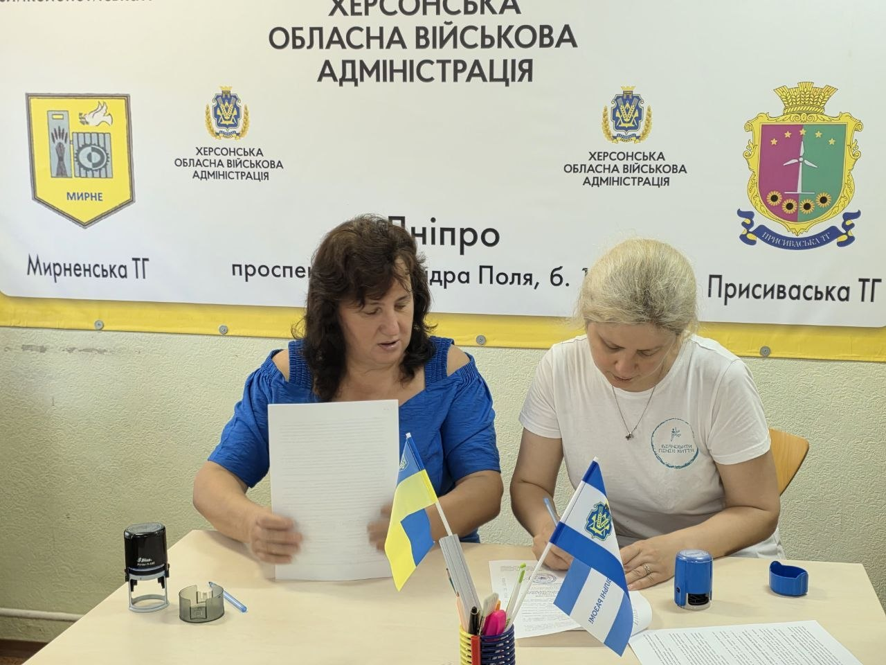 На фото начальниця Мирненської СВА Скадовського району Валентина Головата (ліворуч у синій кофті) та начальниця Присиваської СВА Каховського району Олена Пелешок (праворуч у білій футболці) підписують Меморандум про співпрацю з МБФ «Відновити лінію життя» задля допомоги родинам ВПО. Посадовиці працюють за столом із документами, печатками та прапорцями на тлі банера з символікою Херсонської ОВА, Мирненської та Присиваської громад.