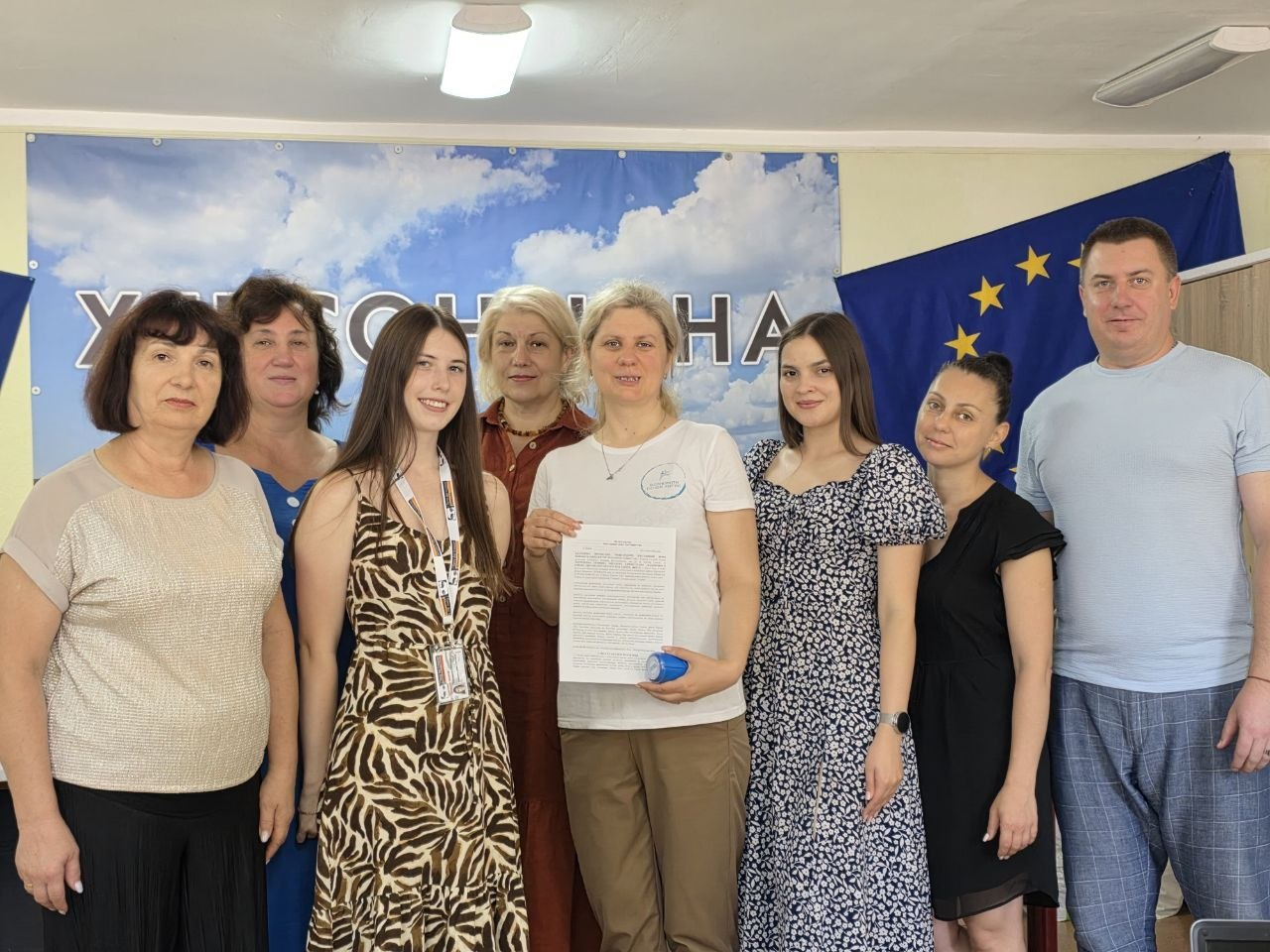 На фото представники Мирненської та Присиваської селищних військових адміністрацій разом із командою МБФ «Відновити лінію життя» після підписання Меморандуму про співпрацю задля допомоги родинам ВПО з Херсонщини. У центрі кадру начальниця Присиваської СВА Олена Пелешок у білій футболці демонструє підписаний документ і печатку, а поруч із нею стоять начальниця Мирненської СВА Валентина Головата та інші учасники зустрічі на тлі прапора Європейського Союзу та банера з написом «Херсонщина».