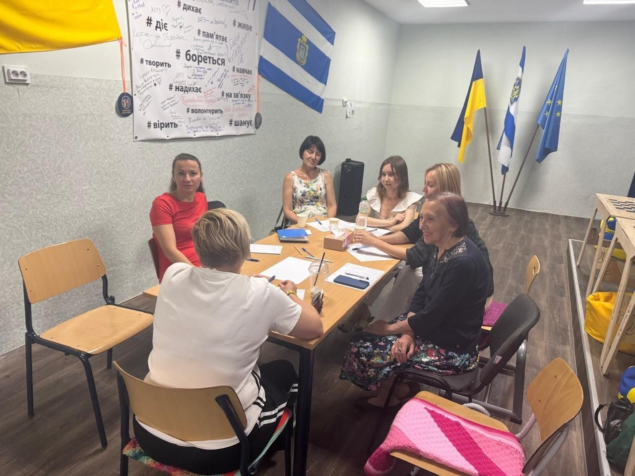 На фото учасники чергової зустрічі групи психологічної підтримки для переселенців із Херсонщини спілкуються за столом у хмельницькому координаційному центрі «Вільні разом». Шість жінок ведуть діалог у кімнаті, прикрашеній прапорами України, Херсонської області та Європейського Союзу, а на стіні позаду них нанесено патріотичні хештеги з центральним написом «# бореться».
