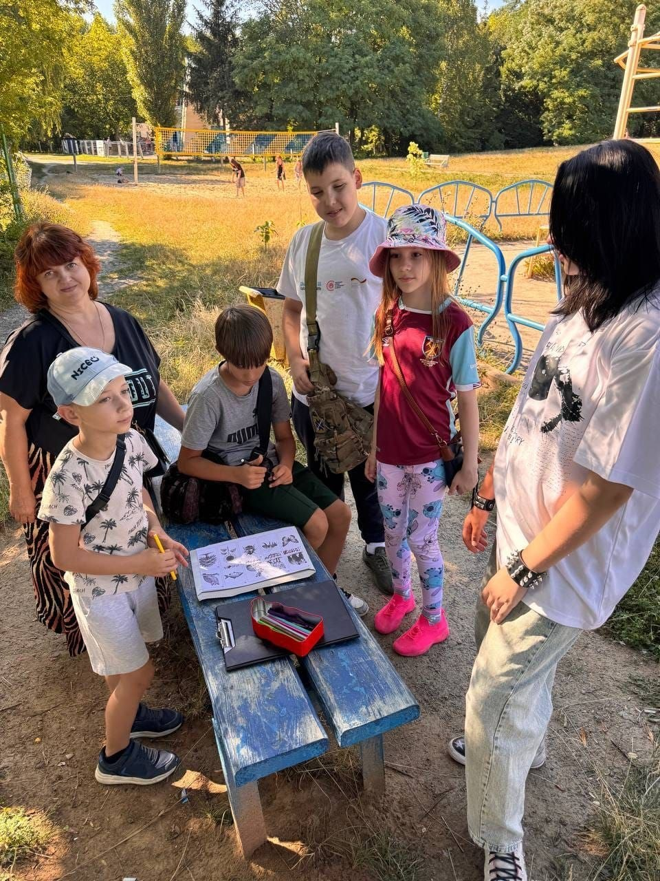 На фото учасники дитячого квесту «Сафарі. Знавці природи», який відбувся у вінницькому креативному просторі «Ми разом!». Група юних переселенців із Херсонщини разом із дорослою наставницею зібралася на вулиці біля синьої дерев'яної лавки, на якій лежать пенал і розгорнута книга з ілюстраціями рослин, на тлі паркової зони та дитячого майданчика.