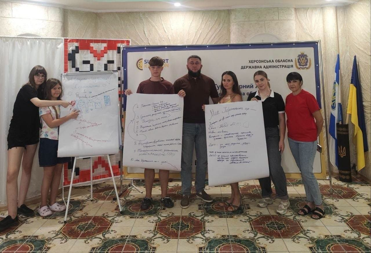 На фото учасники молодіжного флешмобу в Одесі презентують власні візуальні стратегії «Модель розвитку Херсонщини» на найближче десятиліття, розроблені в межах ініціатив Молодіжного конгресу. Сім учасників заходу віком від 14 до 35 років вишикувалися в ряд у приміщенні з символікою Херсонської ОДА: ліворуч дві дівчини показують на мольберт із записами, по центру троє людей тримають великий плакат із тезами стратегії, а праворуч ще дві учасниці демонструють аркуш із написом «Моя Херсонщина це...»