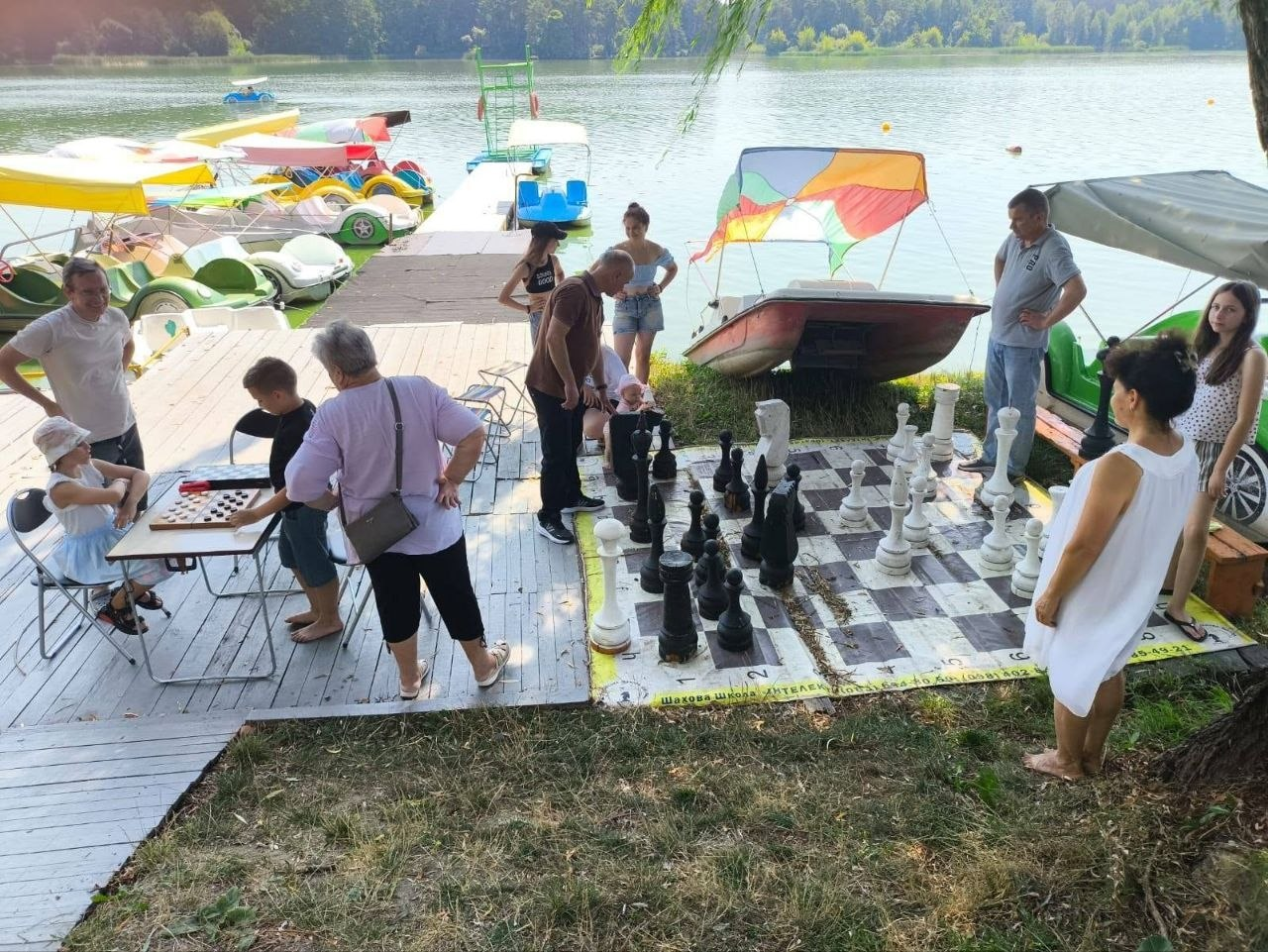 На фото учасники спортивно-дозвіллєвого дня для переселенців із Херсонщини грають у шахи на березі водойми в Житомирі в межах ініціатив Молодіжного конгресу. Ліворуч на дерев'яному причалі за столом триває партія у звичайні настільні шахи, а поруч на траві облаштовано велике підлогове шахове поле, де дорослі та діти переставляють великі чорні й білі фігури на тлі пришвартованих човнів та катамаранів.