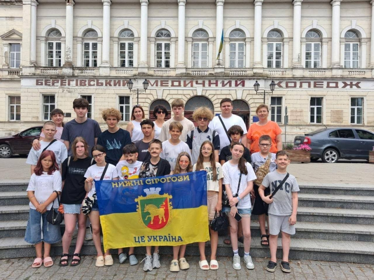 На фото учасники проєкту «Пліч-о-пліч: згуртовані громади» з Нижньосірогозької громади перед будівлею Берегівського медичного коледжу на Закарпатті. Велика група дітей та супроводжуючих дорослих стоїть на сходах історичної споруди, а попереду вони тримають розгорнутий синьо-жовтий прапор із гербом селища Нижні Сірогози та великим написом «НИЖНІ СІРОГОЗИ ЦЕ УКРАЇНА».