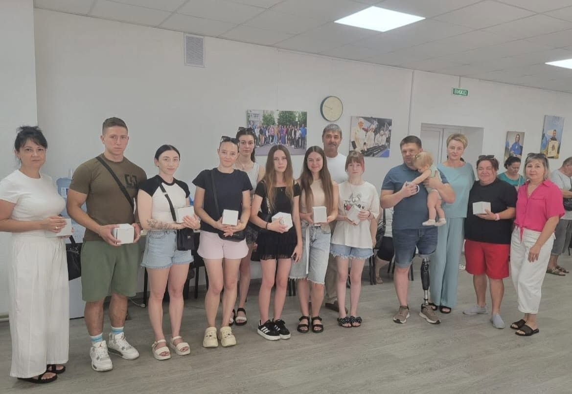 На фото молоді військовослужбовці, ветерани та учнівська молодь Станіславської громади стоять у світлій залі під час відзначення за захист України й активну громадянську позицію. Присутні тримають у руках невеликі білі квадратні коробки з нагородами, а серед нагороджених виділяється чоловік, який тримає на руках маленьку дитину.