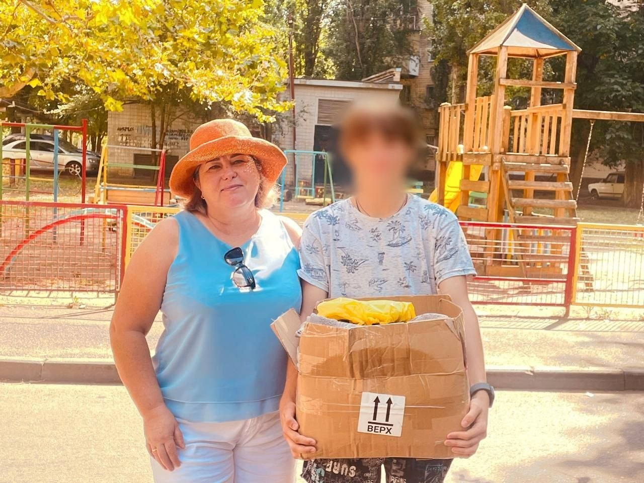 На фото представниця Чаплинської селищної ради стоїть поруч із молодою людиною у світлій футболці, у якої розмите для конфіденційності обличчя та яка тримає велику картонну коробку з гуманітарною допомогою для родин, повернутих з ТОТ. Жінка в помаранчевому капелюсі та блакитному топі посміхається, а на задньому плані видно зелені дерева та дерев'яний дитячий майданчик.