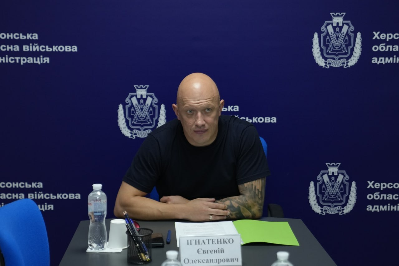 На фото заступник начальника Херсонської ОВА Євгеній Ігнатенко під час виступу на засіданні Урядового комітету з питань гуманітарної та соціальної політики. Чоловік у чорній футболці з татуюваннями на лівій руці сидить за робочим столом із однойменною табличкою на тлі темно-синього банера з емблемами та написами «Херсонська обласна військова адміністрація».