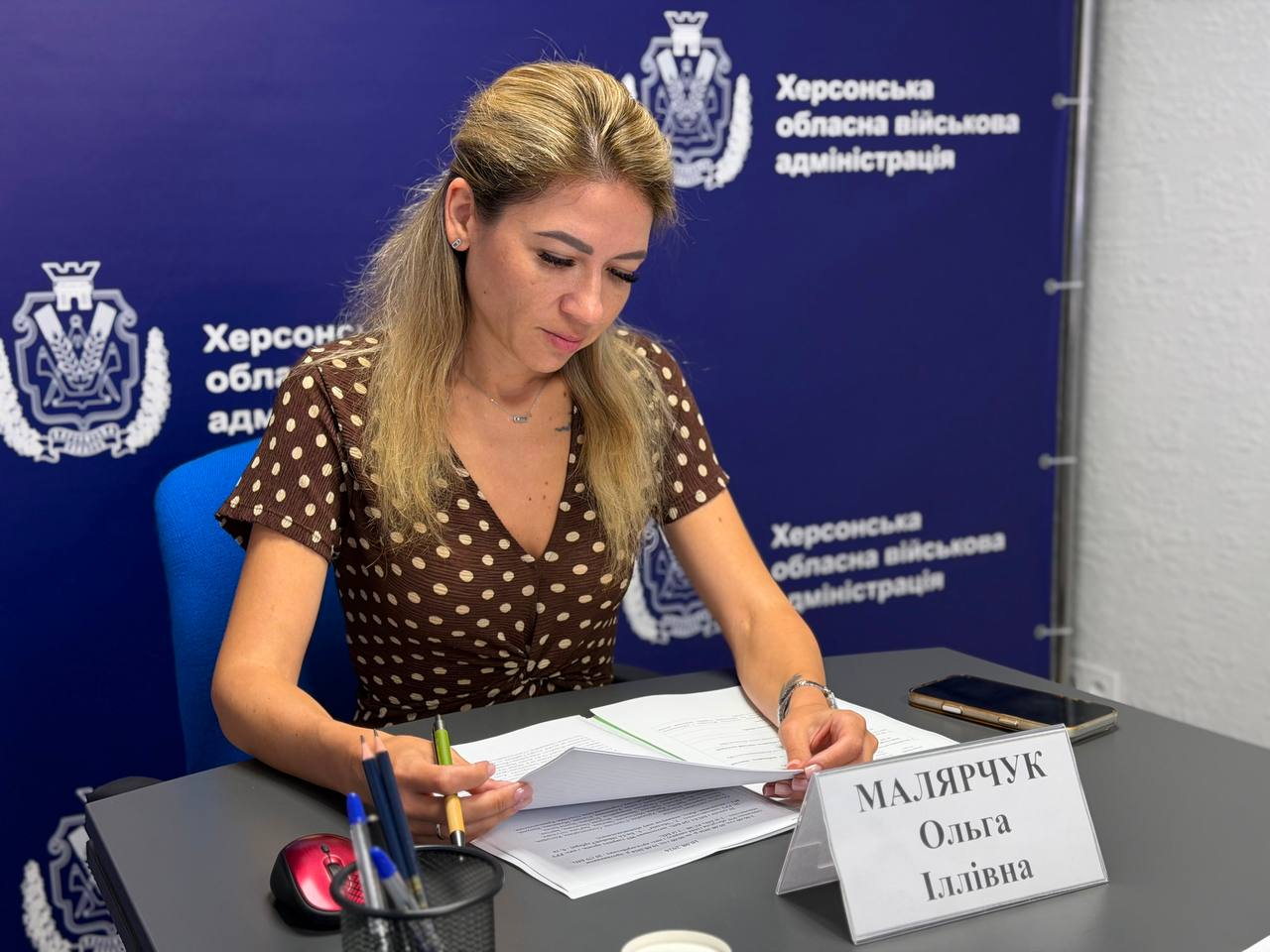 На фото заступниця начальника Херсонської ОВА Ольга Малярчук працює з документами за робочим столом. Жінка в коричневій сукні у білий горошок схилилася над паперами, тримаючи в руці ручку. Перед нею стоїть іменна табличка «МАЛЯРЧУК Ольга Іллівна», а позаду розміщено темно-синій банер із гербами та написами «Херсонська обласна військова адміністрація».