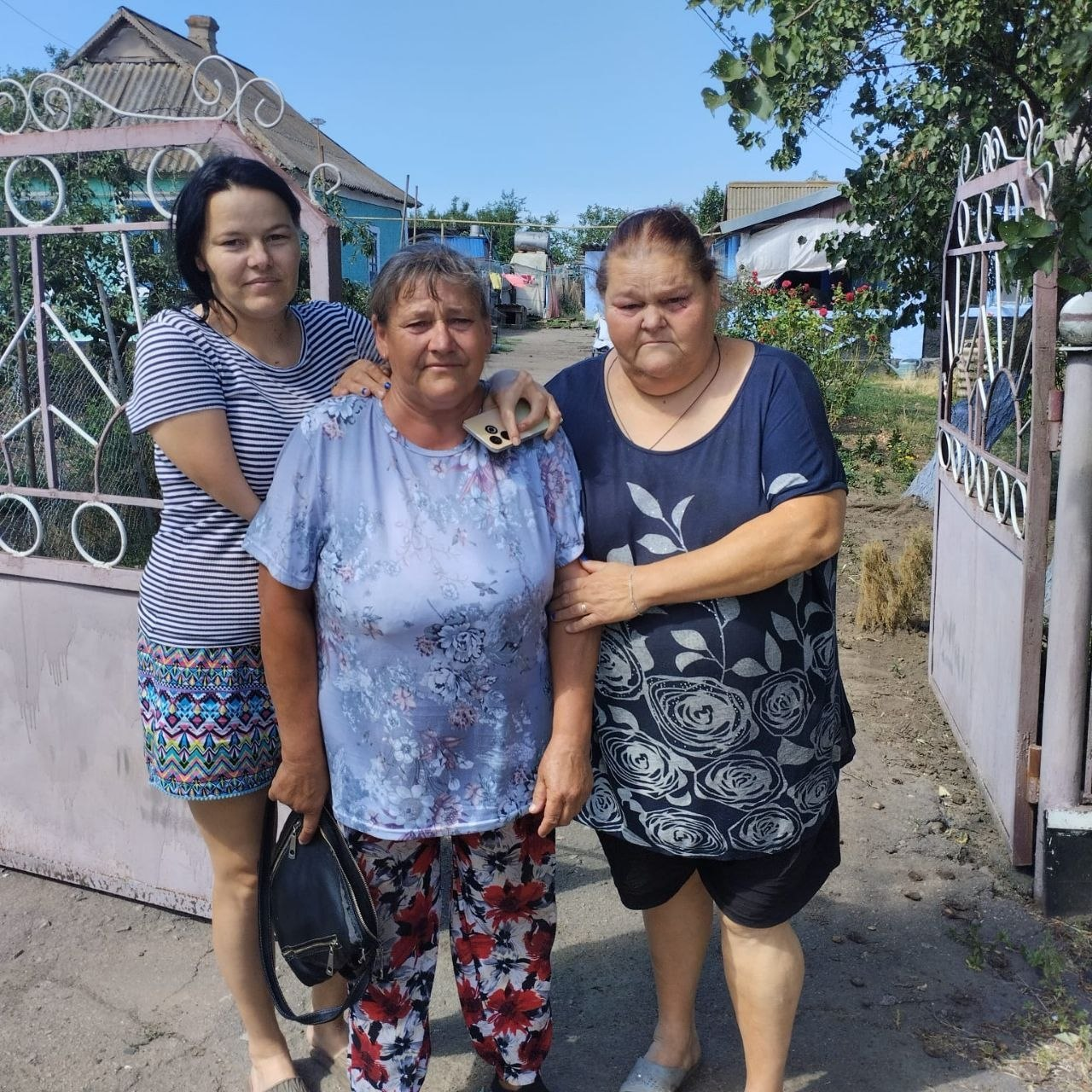 На фото евакуйована жителька села Томарине Бериславської громади (у центрі, в світлій квітчастій блузі та штанах із червоно-чорним візерунком) стоїть в оточенні рідних жінок біля воріт приватного будинку перед виїздом у безпечніші умови. Ліворуч її обіймає за плече молодша жінка у смугастій футболці, а праворуч стоїть жінка в темно-синій блузі з рослинним принтом.