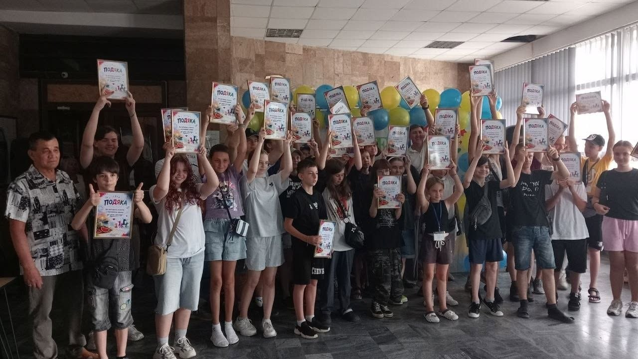 На фото велика група школярів, підлітків та супроводжуючих дорослих стоїть у просторому холі будівлі, тримаючи над головами та перед собою численні офіційні грамоти з написом «ПОДЯКА» за участь в ініціативах Молодіжного конгресу. Учасники дослідження «Мрії та майбутнє молоді Херсонщини» радісно позують для спільного знімка на тлі арки з жовто-блакитних повітряних кульок.