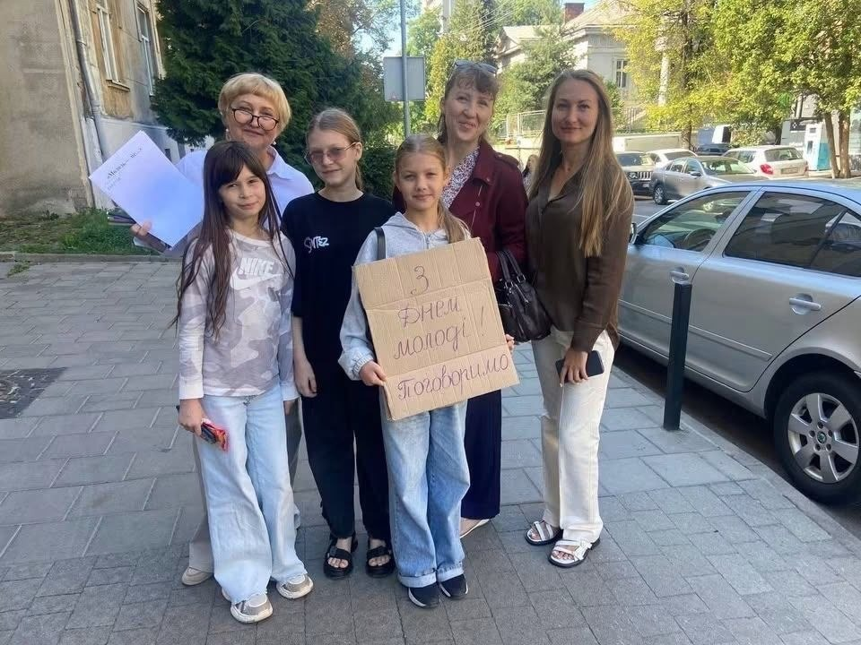 Учасники акції «З Днем молоді. Поговоримо?» від координаційного центру «Вільні разом» у Львові. У центрі — дівчинка з картонним плакатом «З Днем молоді! Поговоримо», поруч стоять молодь та старше покоління переселенців з Херсонщини на фоні міської вулиці.