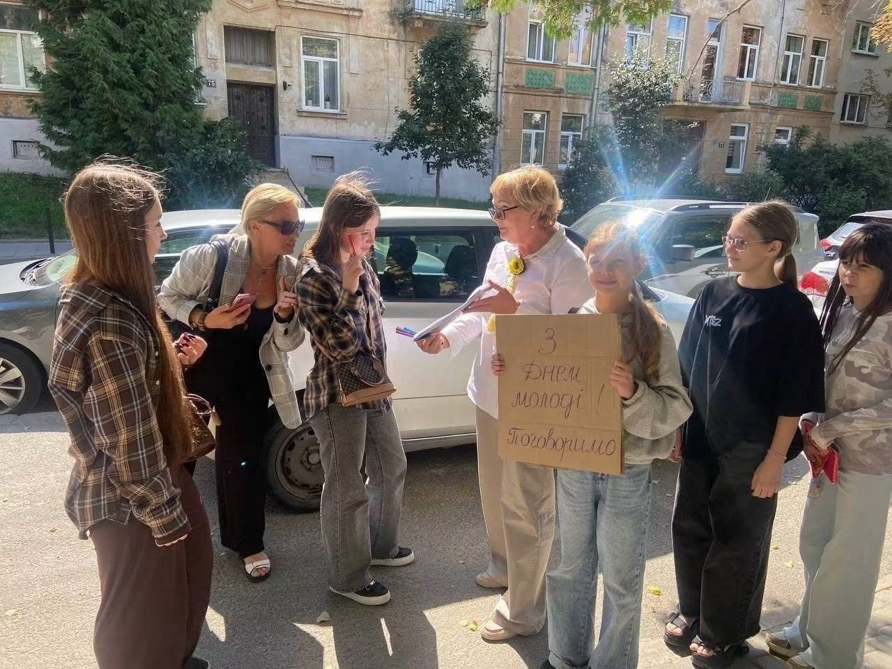 Переселенці з Херсонщини різного віку спілкуються на вулиці Львова під час акції від координаційного центру «Вільні разом». У центрі кола стоїть дівчинка з картонним плакатом «З Днем молоді! Поговоримо». Учасники заходу ведуть живий діалог на тлі припаркованих автомобілів та житлового будинку.