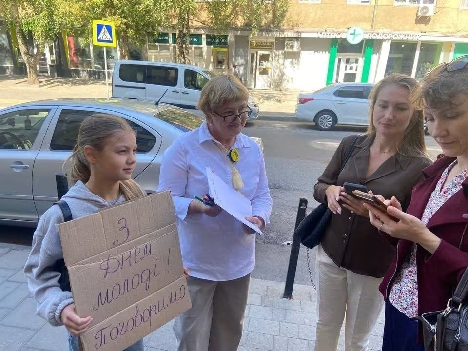 Учасники акції від координаційного центру «Вільні разом» на вулиці Львова. Ліворуч дівчинка тримає плакат «З Днем молоді! Поговоримо», поруч стоїть жінка з паперами, а дві інші жінки дивляться в екран смартфона. На задньому плані видно припарковані авто, аптеку «Подорожник» та Ощадбанк.