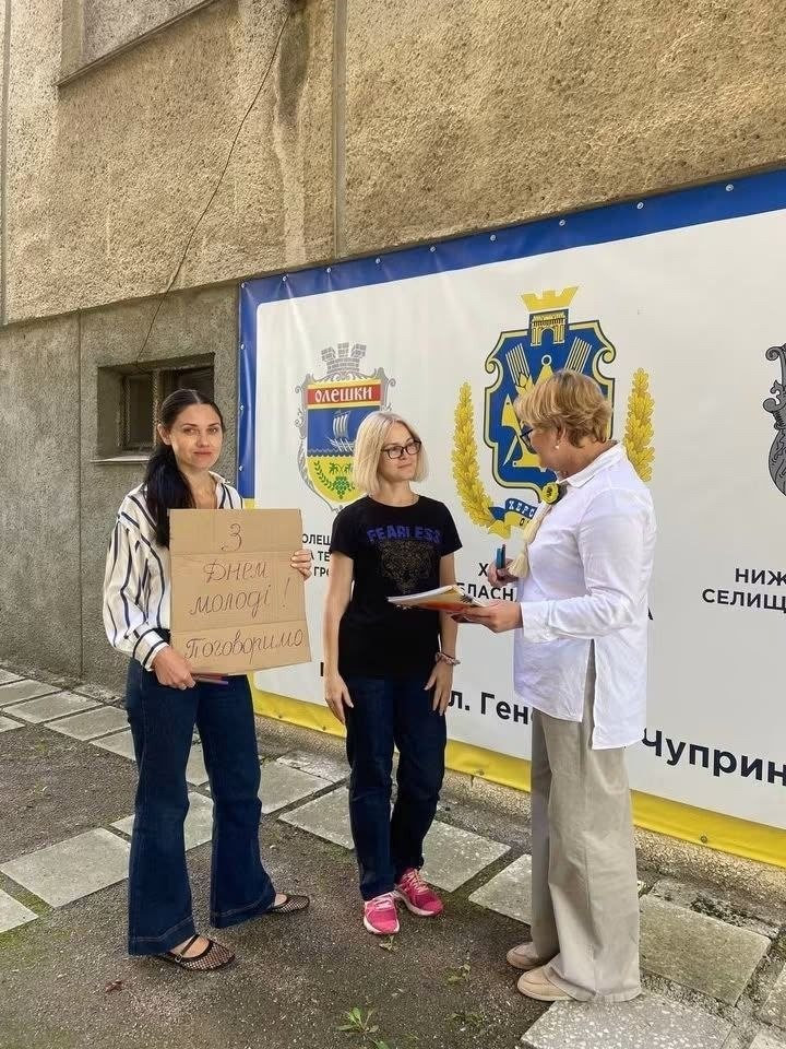 Три учасниці акції від координаційного центру «Вільні разом» стоять біля будівлі у Львові. Ліворуч дівчина тримає картонний плакат «З Днем молоді! Поговоримо». Поруч стоїть дівчина у чорній футболці, а жінка у білій блузі тримає папери. Вони знаходяться на тлі банера з гербами Олешок та Херсонщини і написом «вул. Генерала Чупринки».