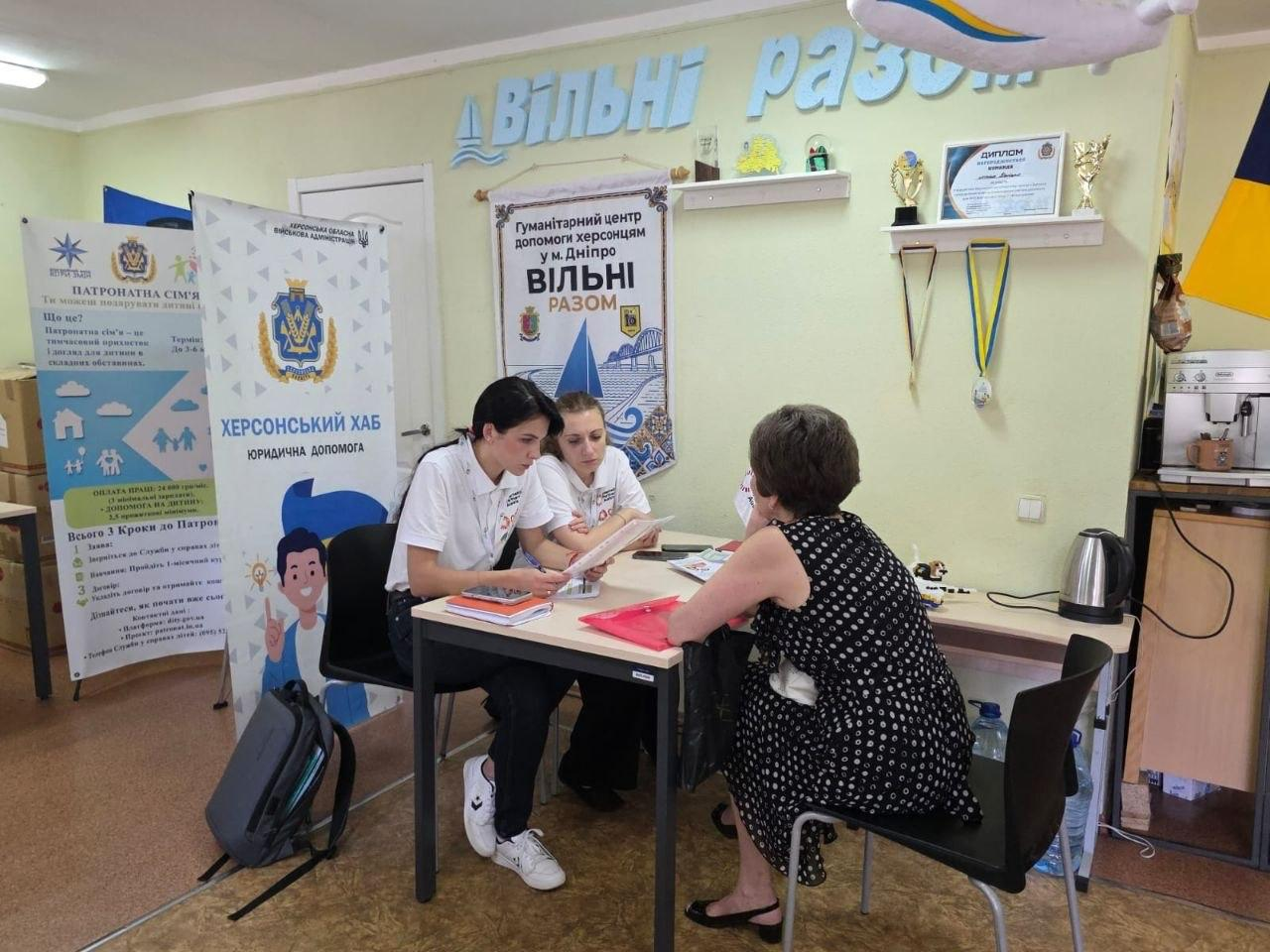 Консультація переселенки з Херсонщини в гуманітарному центрі «Вільні разом» у Дніпрі. Юрисконсульт ГО «Аваліст» та кейс-менеджер у білих футболках сидять за столом і вивчають документи разом із відвідувачкою. На стіні за ними розміщено банер «Гуманітарний центр допомоги херсонцям у м. Дніпро „Вільні разом“».